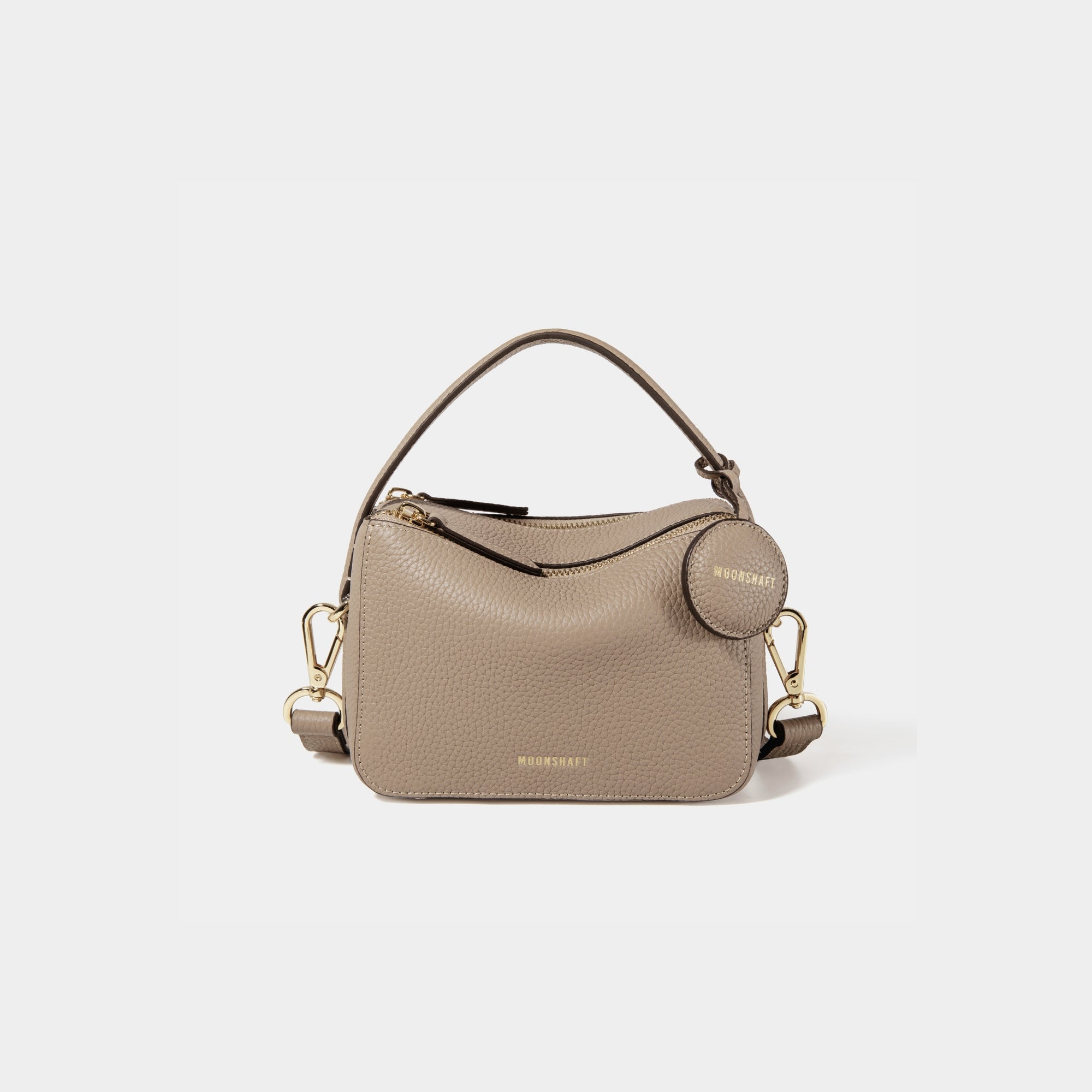 A close-up of the Mini Double Zip Bag in a sandstone color, made from pebbled leather. The bag features a top handle and an adjustable shoulder strap with gold-tone hardware. It has two zippered compartments for storage and a round leather tag attached to the side, embossed with the brand logo 'MOONSHAFT' in gold. The minimalist and structured design enhances its modern and stylish appeal.