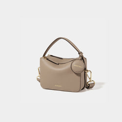A side-front view of the Mini Double Zip Bag in sandstone color, made from pebbled leather. The bag features a top handle, gold-tone hardware, and an adjustable shoulder strap. It has two zippered compartments and a small round leather tag attached to the side, embossed with the 'MOONSHAFT' logo in gold. The clean and structured design showcases its minimalist elegance and functional style.
