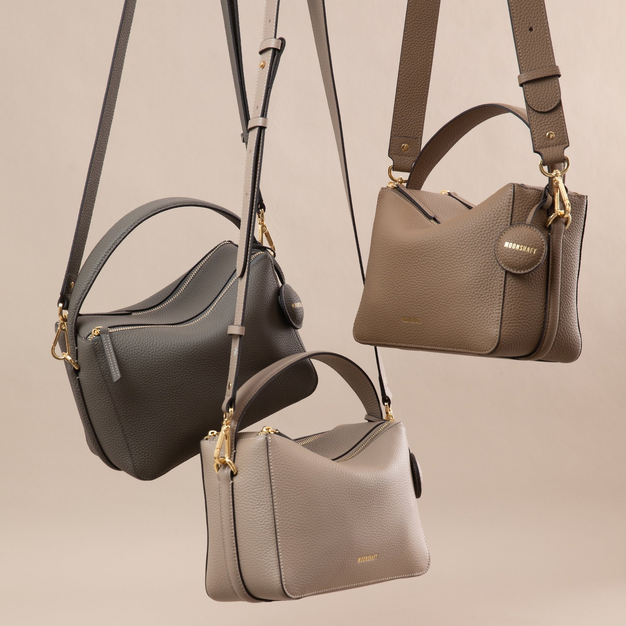 Three MOONSHAFT upgraded double zip pebbled leather bags in neutral tones (charcoal, driftwood, and sandstone) are stylishly suspended by their straps. Each bag showcases a structured design with gold-tone zippers and hardware, along with a top handle and an adjustable shoulder strap. The smooth pebbled leather texture and minimalist aesthetic highlight the elegance and versatility of the bags, making them perfect for various outfits and occasions. The neutral background complements the sophisticated color