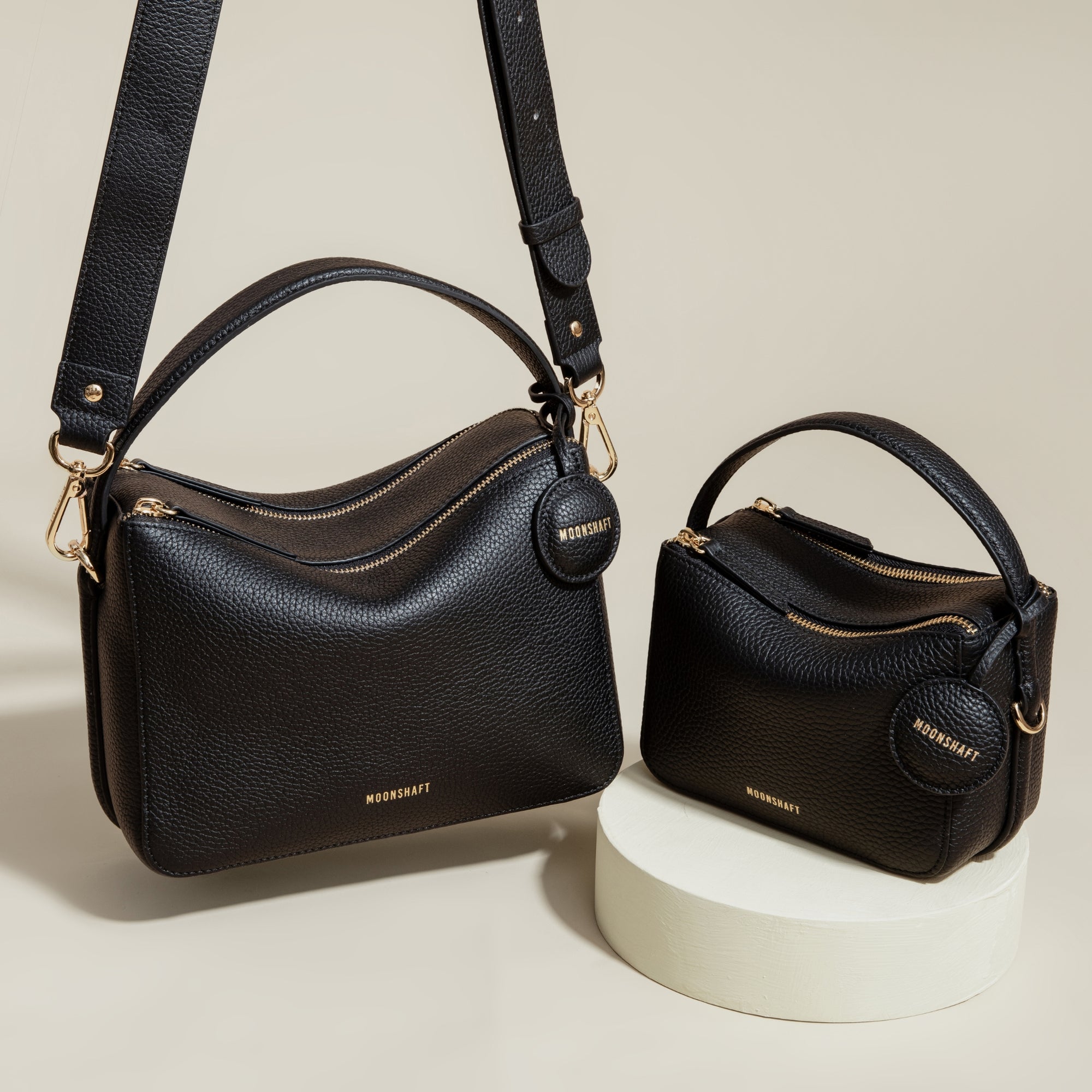 A side-by-side display of two black MOONSHAFT bags: the Double Zip Bag and the smaller Mini Double Zip Bag in black. Both bags are made from pebbled leather, featuring gold-tone hardware, zippered compartments, and round leather tags embossed with the 'MOONSHAFT' logo. The structured and minimalist design of both bags highlights their modern, elegant aesthetic