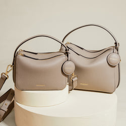 A side-by-side display of two sandstone-colored MOONSHAFT bags: the larger Original Double Zip Bag and the smaller Mini Double Zip Bag. Both bags are made from pebbled leather and feature gold-tone hardware, zippered compartments, and round leather tags embossed with the 'MOONSHAFT' logo. The structured, minimalist design highlights the sophisticated and timeless aesthetic of both bags.