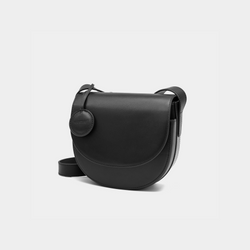 A front-side angle view of a black half-moon crossbody bag made of smooth leather. The bag has a curved base and minimalistic flap closure. A circular leather tag is attached to the strap, adding a subtle design detail. The adjustable strap and sleek design offer a versatile, elegant look.