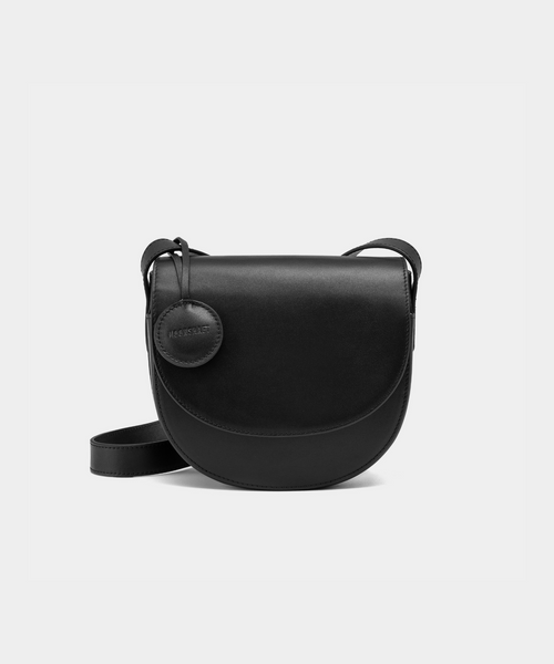 A black half-moon crossbody bag with a smooth leather finish, featuring a circular leather tag attached to the strap. The structured design highlights the bag's minimalist aesthetic and sleek curves, offering an elegant yet functional style for everyday wear.