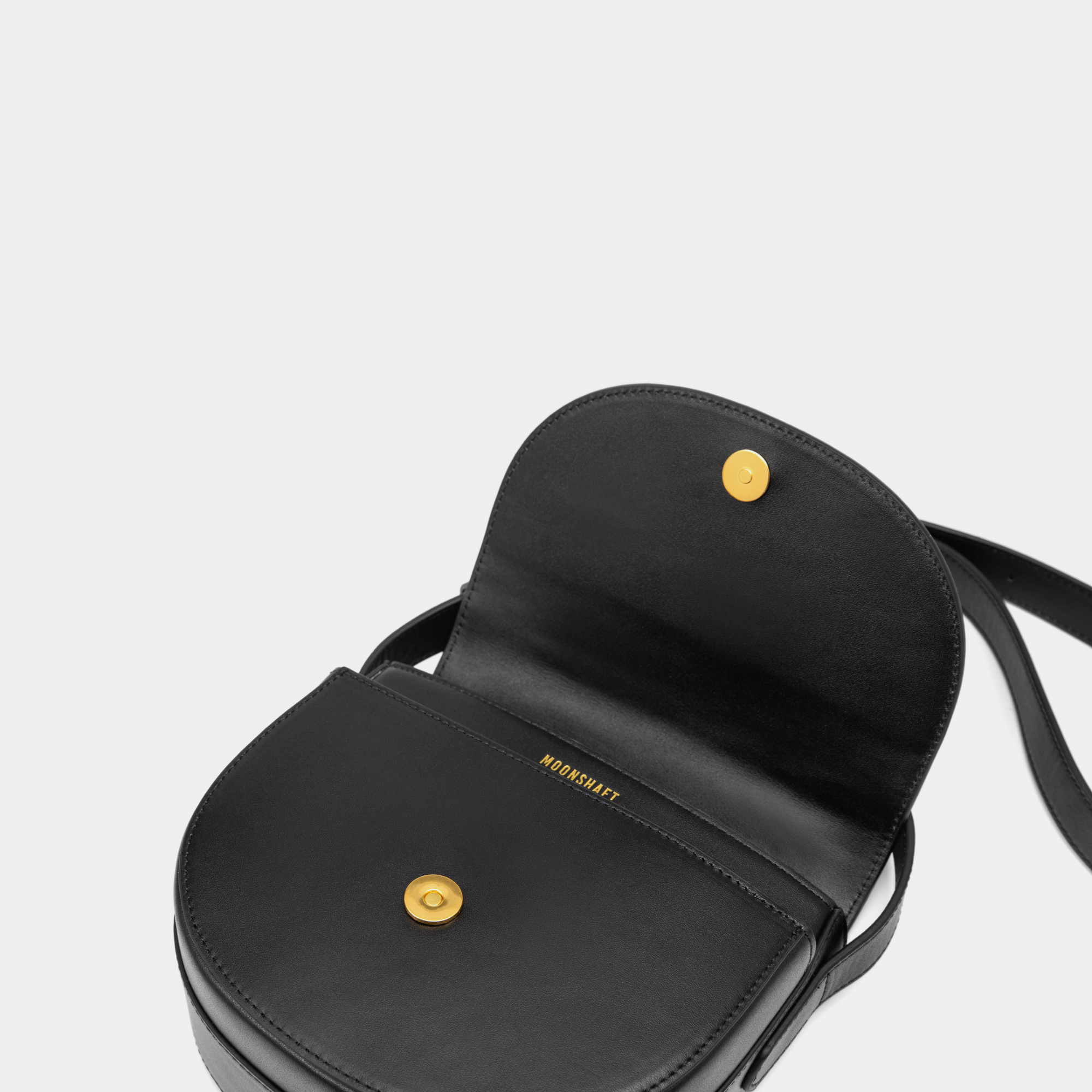 The close-up view displays the interior of the half-moon crossbody bag with its smooth leather flap open. A gold-tone magnetic closure is prominently featured, offering a secure and stylish fastening. The inner lining is minimalistic, with the brand name "MOONSHAFT" subtly embossed in gold just beneath the closure for a refined touch.