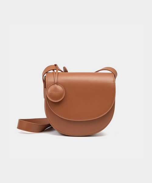 A caramel half-moon crossbody bag with a smooth leather finish, featuring a circular leather tag attached to the strap. The structured design highlights the bag's minimalist aesthetic and sleek curves, offering an elegant yet functional style for everyday wear.