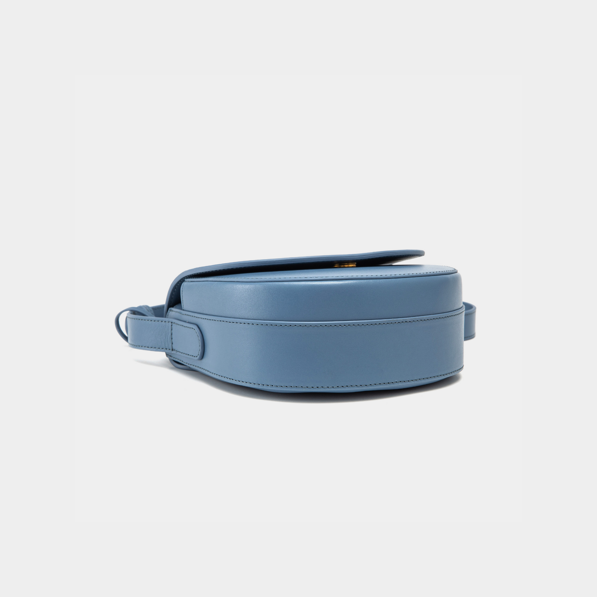 A bottom view of the half-moon crossbody bag in stone blue leather, showcasing its structured base. The smooth and clean design is accentuated by minimal stitching along the edges, ensuring durability and reinforcing the bag's sleek silhouette. The strap attaches securely to the sides of the base, maintaining the overall elegance of the bag.