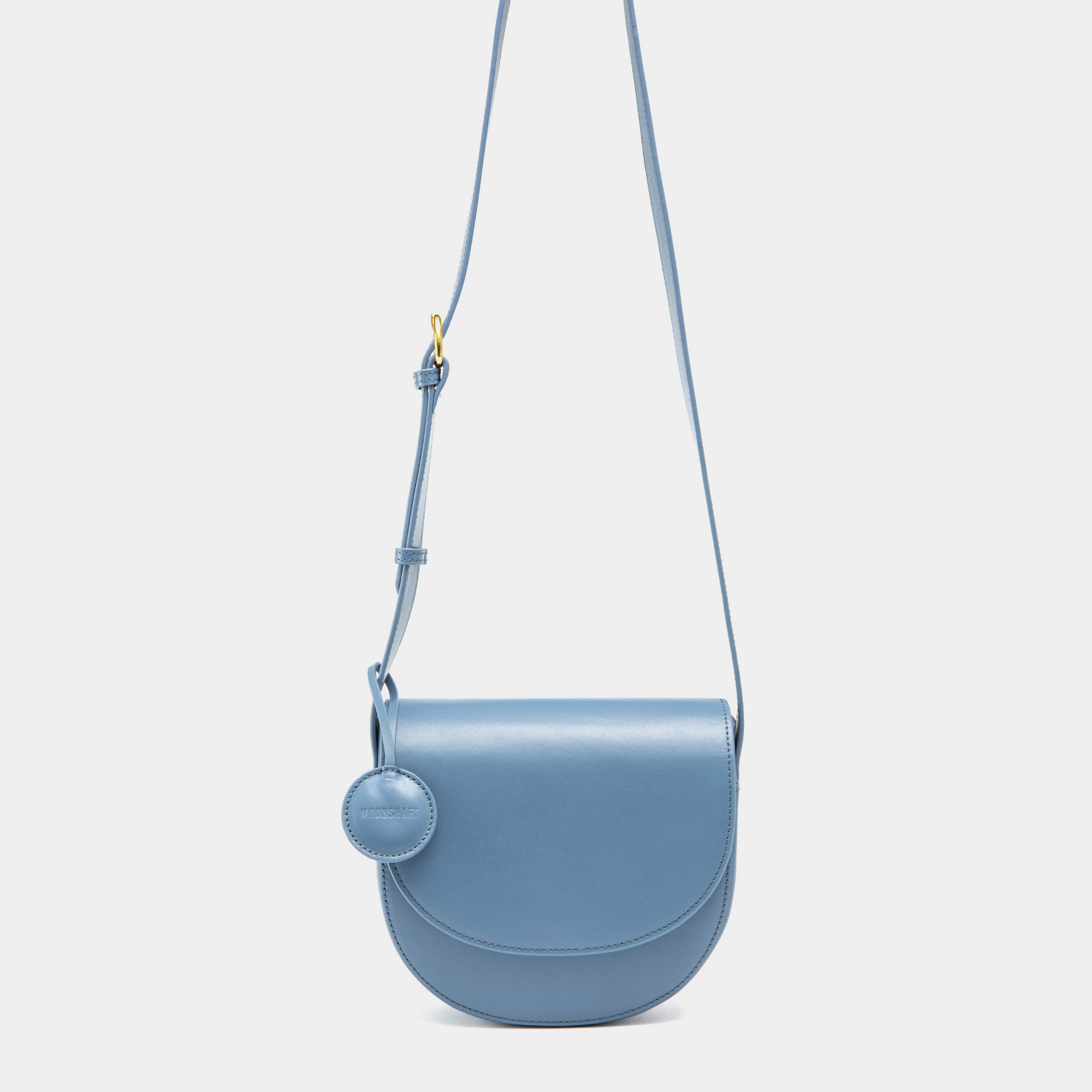 A sleek black half-moon crossbody bag with a curved, structured silhouette. The bag has a smooth leather finish and features a circular leather tag attached to the strap. The adjustable leather strap with a gold-tone buckle adds a touch of sophistication to the minimalist design.