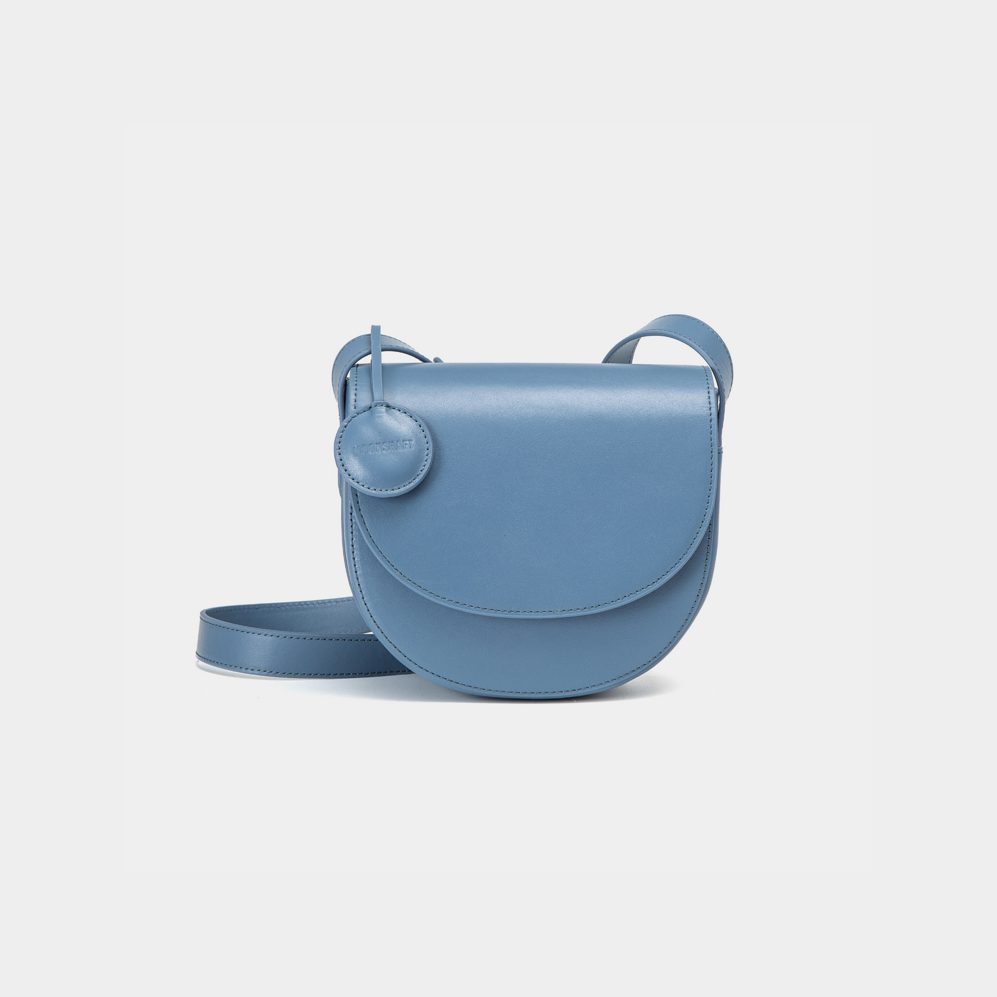 A black half-moon crossbody bag with a smooth leather finish, featuring a circular leather tag attached to the strap. The structured design highlights the bag's minimalist aesthetic and sleek curves, offering an elegant yet functional style for everyday wear.