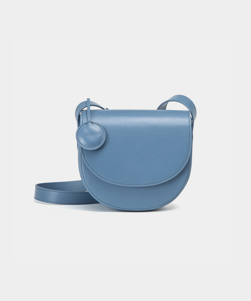 A black half-moon crossbody bag with a smooth leather finish, featuring a circular leather tag attached to the strap. The structured design highlights the bag's minimalist aesthetic and sleek curves, offering an elegant yet functional style for everyday wear.