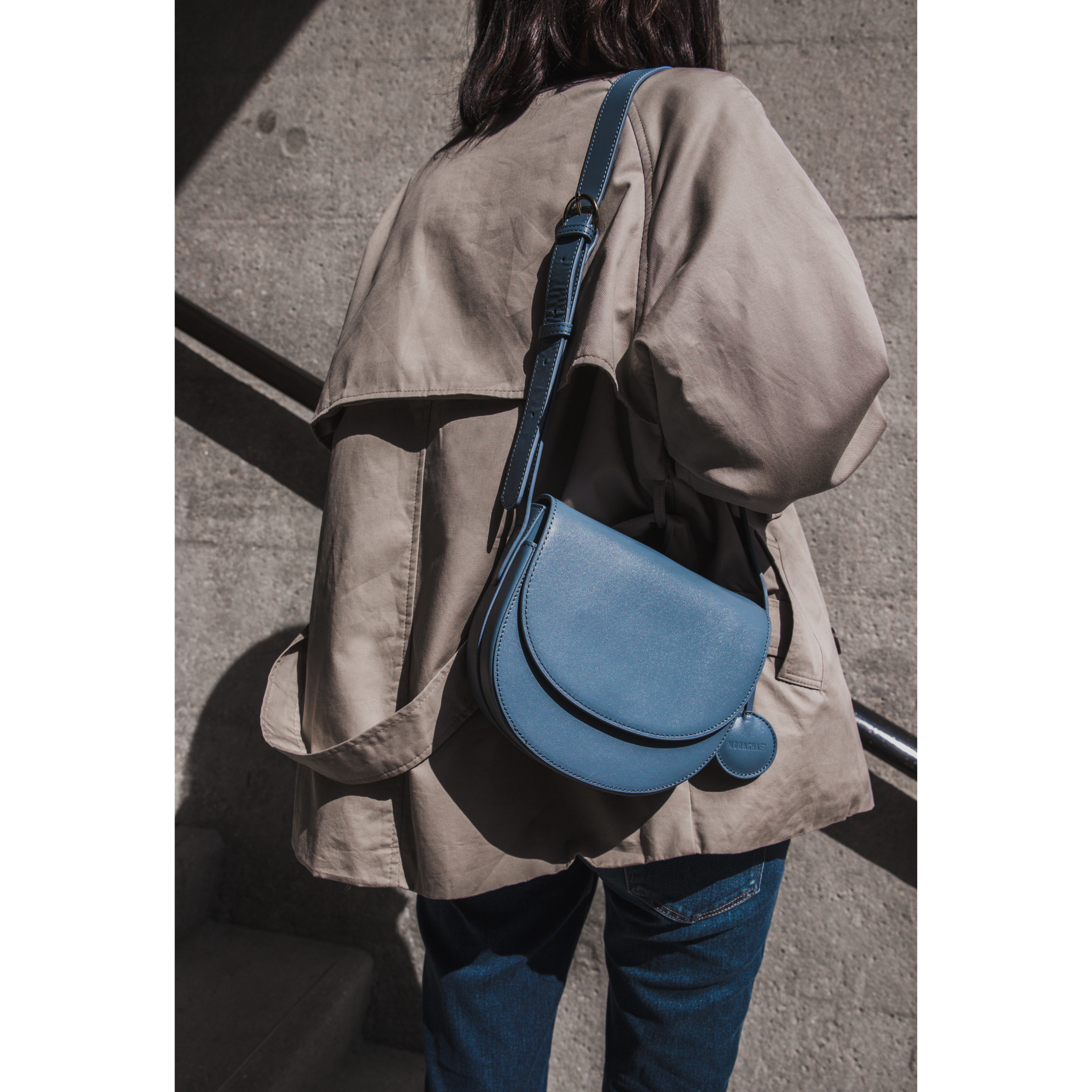 A model walks down a stairwell, wearing a beige jacket and carrying the half-moon crossbody bag in stone blue color over the shoulder. The rich blue of the bag contrasts with the muted neutral tones of the outfit, standing out against the stone background. The circular leather tag complements the bag’s rounded silhouette, while the adjustable strap ensures both comfort and style.