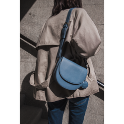A model walks down a stairwell, wearing a beige jacket and carrying the half-moon crossbody bag in stone blue color over the shoulder. The rich blue of the bag contrasts with the muted neutral tones of the outfit, standing out against the stone background. The circular leather tag complements the bag’s rounded silhouette, while the adjustable strap ensures both comfort and style.