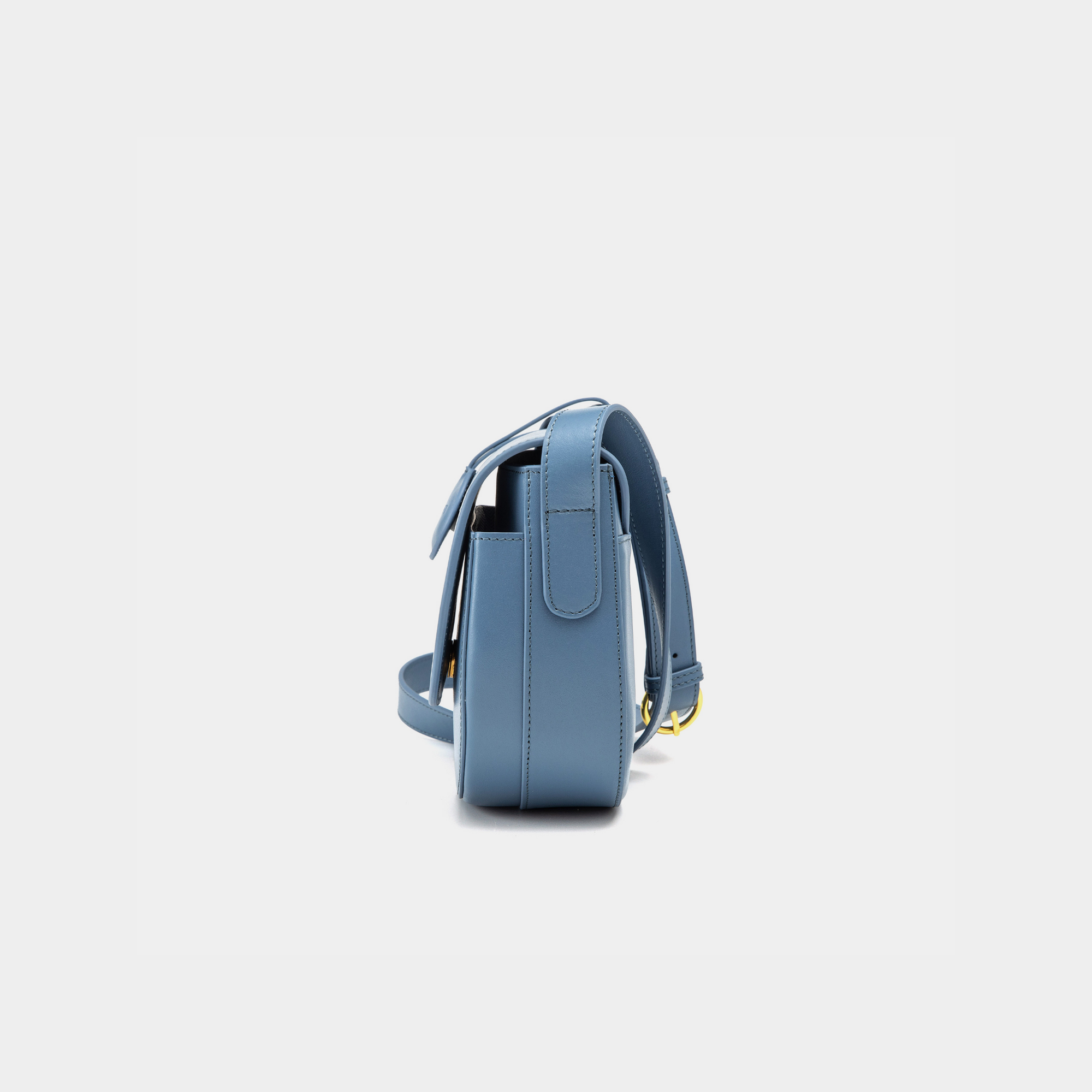 A side view of a stone blue half-moon crossbody bag showcasing its structured profile. The bag has a sleek, smooth leather finish, and its curved base complements the minimalistic design. The adjustable leather strap and gold-tone hardware are visible from this angle, adding a touch of refinement.