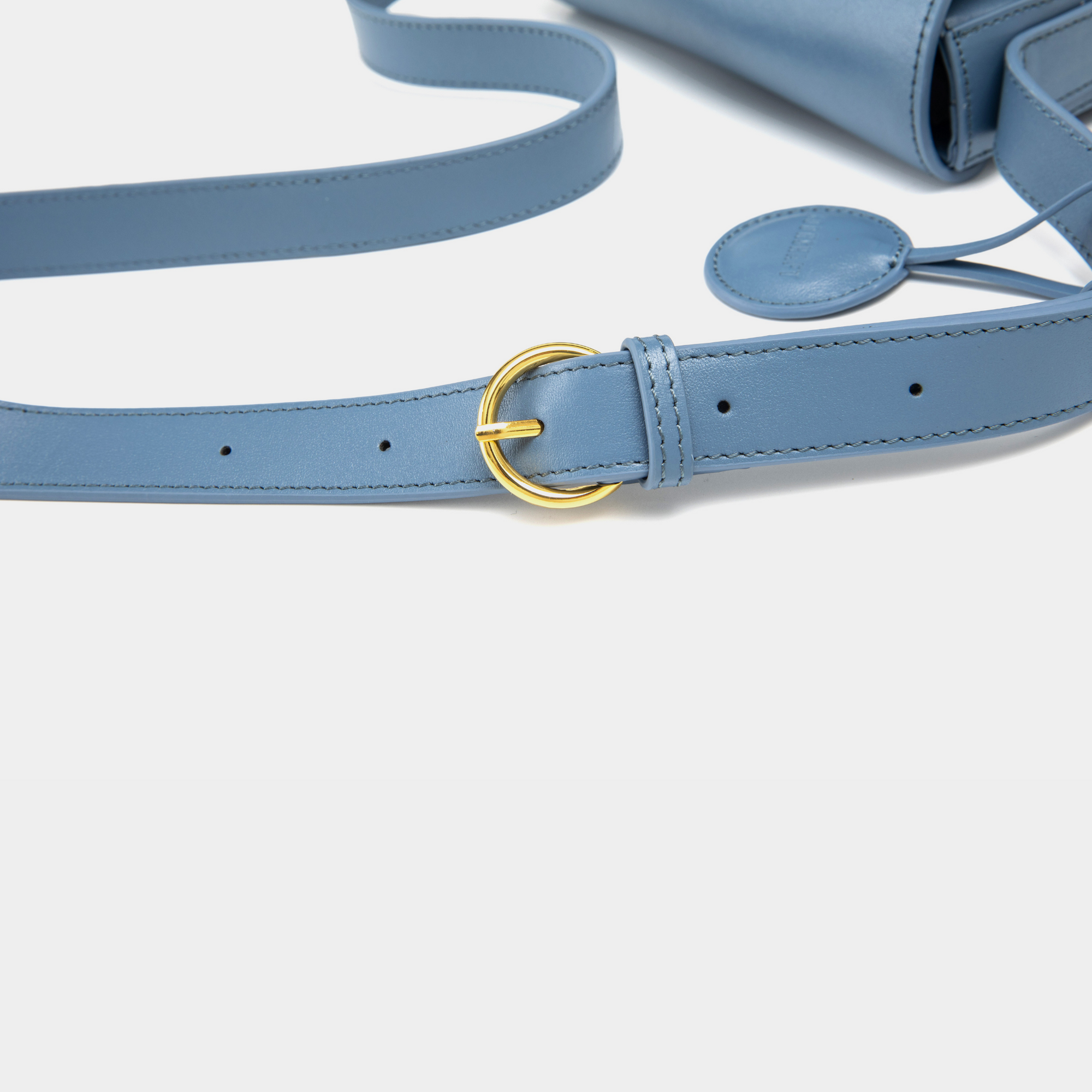 A close-up of a stone blue-colored leather crossbody bag strap, showcasing its adjustable buckle design in polished gold. The stitching is neatly aligned along the edges, emphasizing the quality of craftsmanship. The smooth leather strap lies flat, with the circular leather tag in the background, complementing the overall design of the bag.