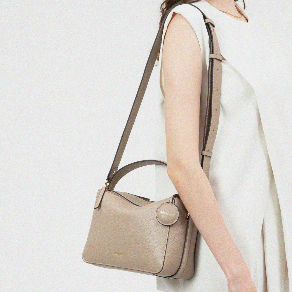 A model wearing a sleeveless cream outfit carries the MOONSHAFT double zip bag in sandstone color over her shoulder. The bag, made from pebbled leather, features a top handle, an adjustable shoulder strap, gold-tone hardware, and a round leather tag embossed with the 'MOONSHAFT' logo. The bag’s structured design and neutral tone complement the model's minimalist, elegant style.