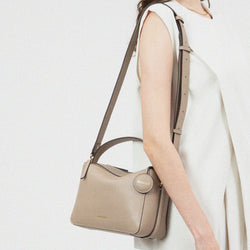 A model wearing a sleeveless cream outfit carries the MOONSHAFT double zip bag in sandstone color over her shoulder. The bag, made from pebbled leather, features a top handle, an adjustable shoulder strap, gold-tone hardware, and a round leather tag embossed with the 'MOONSHAFT' logo. The bag’s structured design and neutral tone complement the model's minimalist, elegant style.