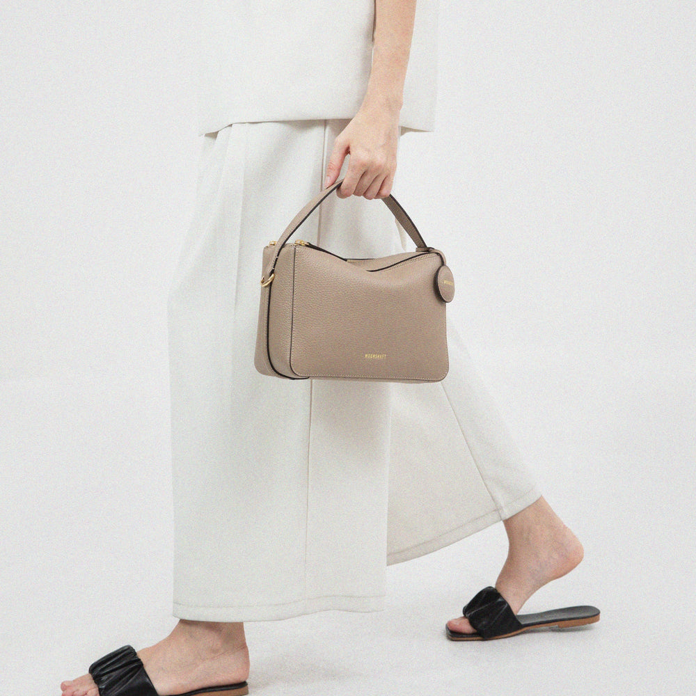 A person dressed in white holding the MOONSHAFT Double Zip Bag in sandstone color. The taupe-colored leather bag features a minimalist design with a structured silhouette and a circular leather tag, paired with gold-tone hardware.