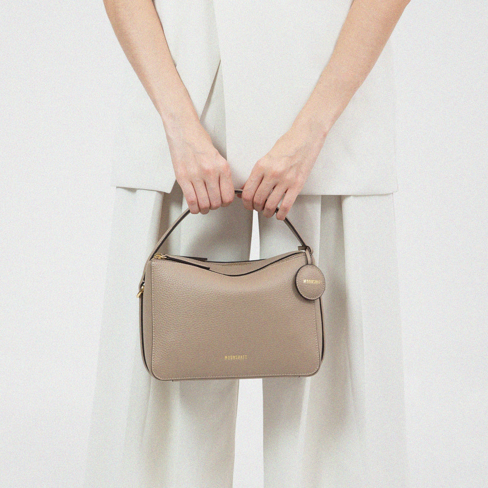 A model in light-colored clothing is holding a sandstone-colored doublr zip bag from MOONSHAFT, featuring a structured design and a circular leather tag with gold-tone hardware