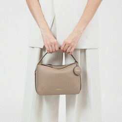 A model in light-colored clothing is holding a sandstone-colored doublr zip bag from MOONSHAFT, featuring a structured design and a circular leather tag with gold-tone hardware