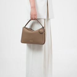 This image features the Double Zip Bag in a muted taupe shade. The model is holding the bag by its top handle, highlighting its structured and elegant form. The bag is made from pebbled leather and comes with gold-tone hardware, including the zippers and a signature round leather tag embossed with the 'MOONSHAFT' logo. The minimalist yet chic design adds sophistication to the overall look, making it a versatile accessory for various occasions.