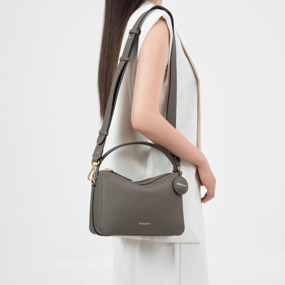 This image showcases the Double Zip Bag in a grayish hue, styled with a structured design and adorned with gold-tone hardware. The model is wearing it crossbody, highlighting its versatility and minimalist appeal. The round leather tag embossed with the 'MOONSHAFT' logo adds a signature touch to the overall sleek and functional look of the bag.