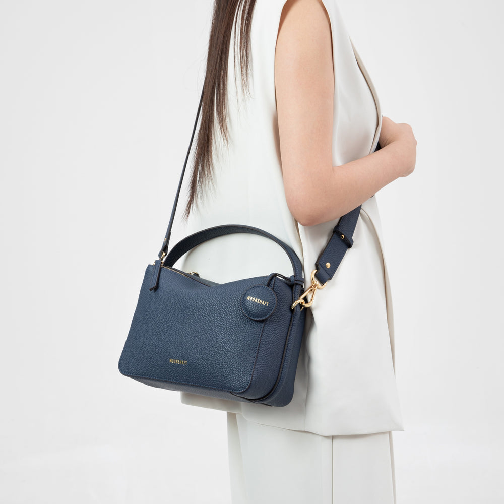 A model dressed in a sleeveless cream outfit carries the MOONSHAFT Double Zip Bag in a marine color. The bag, crafted from pebbled leather, features gold-tone hardware, a top handle, an adjustable shoulder strap, and a round leather tag embossed with the 'MOONSHAFT' logo. The structured design of the bag complements the sleek, modern aesthetic of the model's outfit.