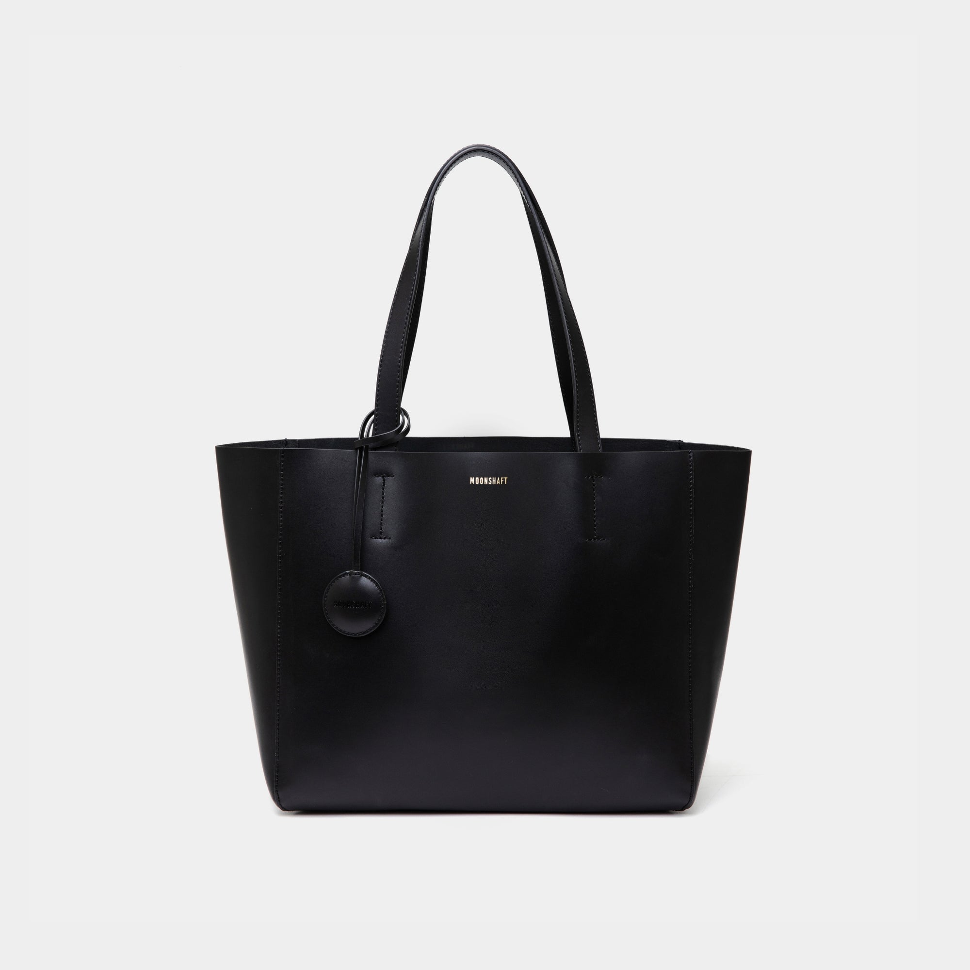 A minimalist black leather tote bag displayed against a plain background. The tote features a smooth leather texture and a clean, structured design with no external embellishments, showcasing its simplicity and elegance. It has two sturdy leather handles and a spacious interior, making it a practical choice for everyday use. The neutral tan color adds to the bag’s versatile appeal, perfect for complementing various outfits and occasions.