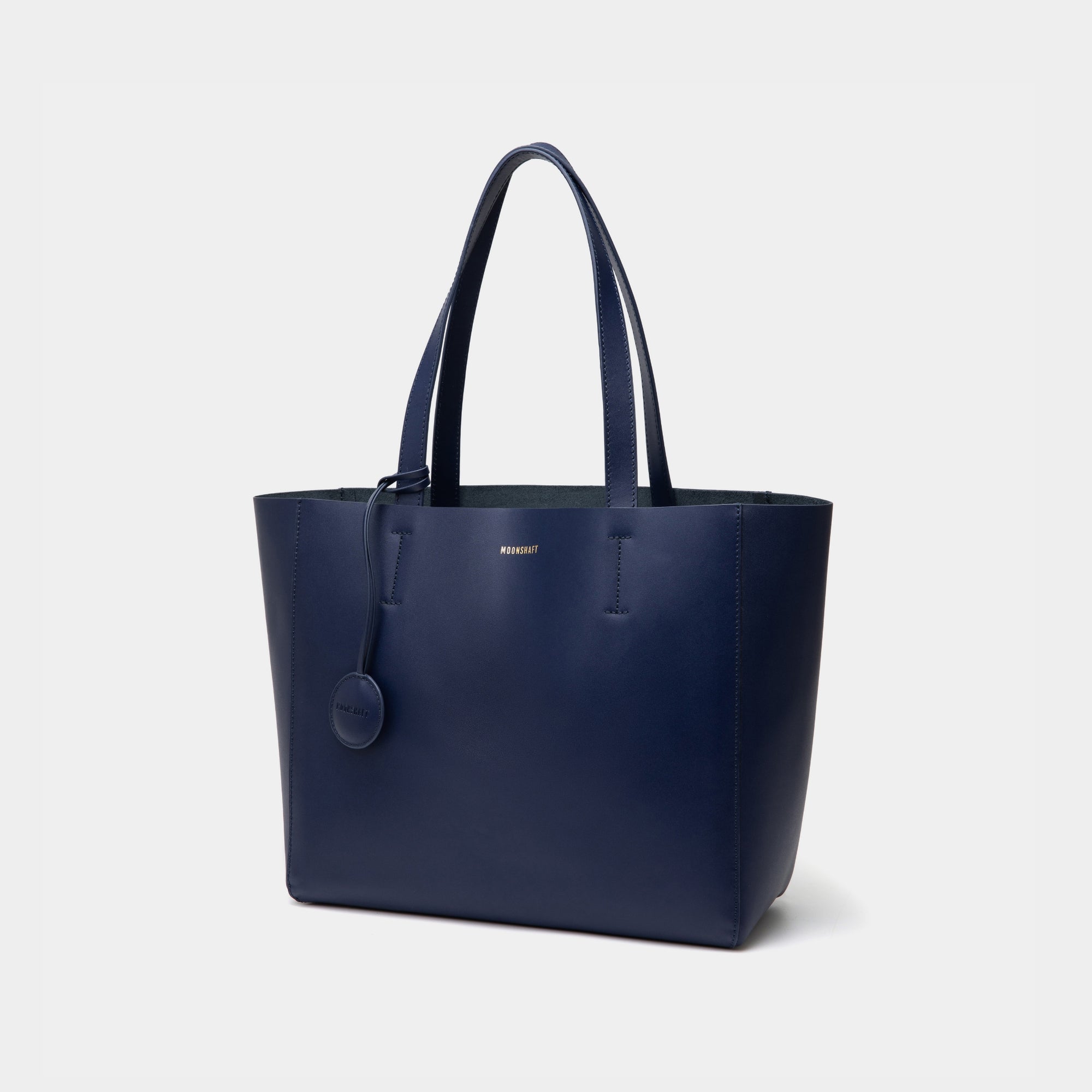 The diagonal view of the navy MOONSHAFT tote bag displayed against a white background. The bag features a sleek, minimalist design with reinforced stitching along the handles, emphasizing its spacious and structured silhouette.