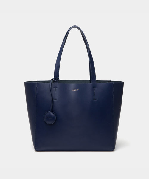 A minimalist navy leather tote bag displayed against a plain background. The tote features a smooth leather texture and a clean, structured design with no external embellishments, showcasing its simplicity and elegance. It has two sturdy leather handles and a spacious interior, making it a practical choice for everyday use. The neutral tan color adds to the bag’s versatile appeal, perfect for complementing various outfits and occasions.