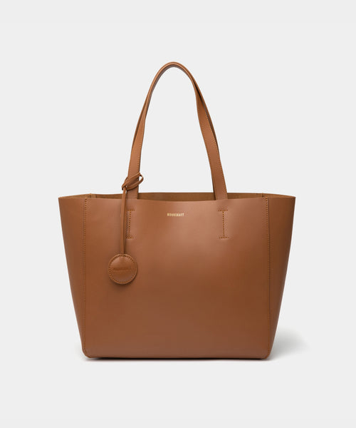 A minimalist caramel leather tote bag displayed against a plain background. The tote features a smooth leather texture and a clean, structured design with no external embellishments, showcasing its simplicity and elegance. It has two sturdy leather handles and a spacious interior, making it a practical choice for everyday use. The neutral tan color adds to the bag’s versatile appeal, perfect for complementing various outfits and occasions.