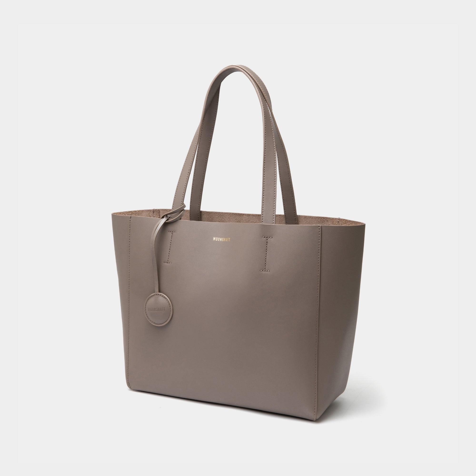 The diagonal view of the taupe MOONSHAFT tote bag displayed against a white background. The bag features a sleek, minimalist design with reinforced stitching along the handles, emphasizing its spacious and structured silhouette.