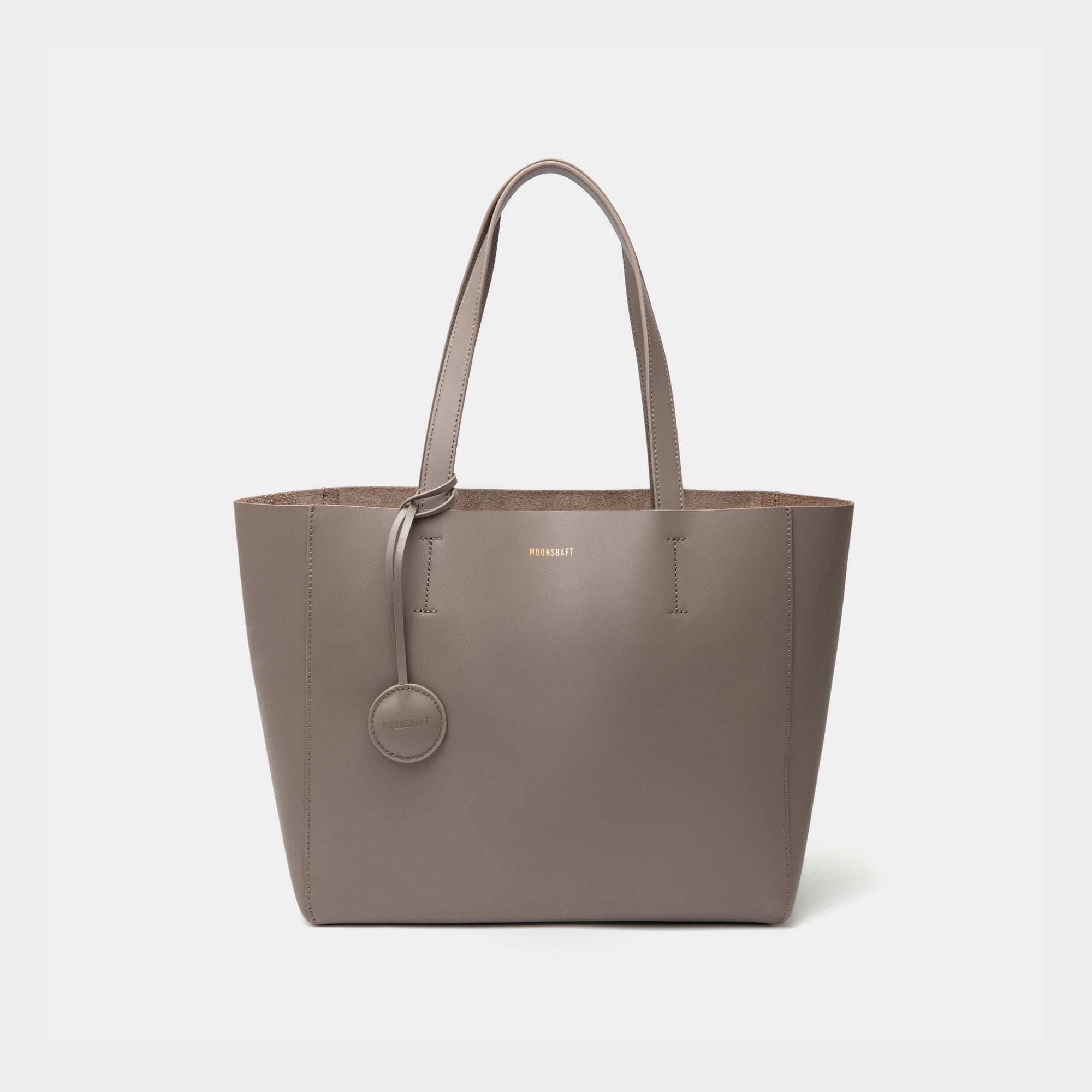 A minimalist taupe leather tote bag displayed against a plain background. The tote features a smooth leather texture and a clean, structured design with no external embellishments, showcasing its simplicity and elegance. It has two sturdy leather handles and a spacious interior, making it a practical choice for everyday use. The neutral tan color adds to the bag’s versatile appeal, perfect for complementing various outfits and occasions.
