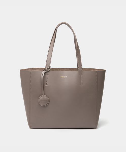 A minimalist taupe leather tote bag displayed against a plain background. The tote features a smooth leather texture and a clean, structured design with no external embellishments, showcasing its simplicity and elegance. It has two sturdy leather handles and a spacious interior, making it a practical choice for everyday use. The neutral tan color adds to the bag’s versatile appeal, perfect for complementing various outfits and occasions.