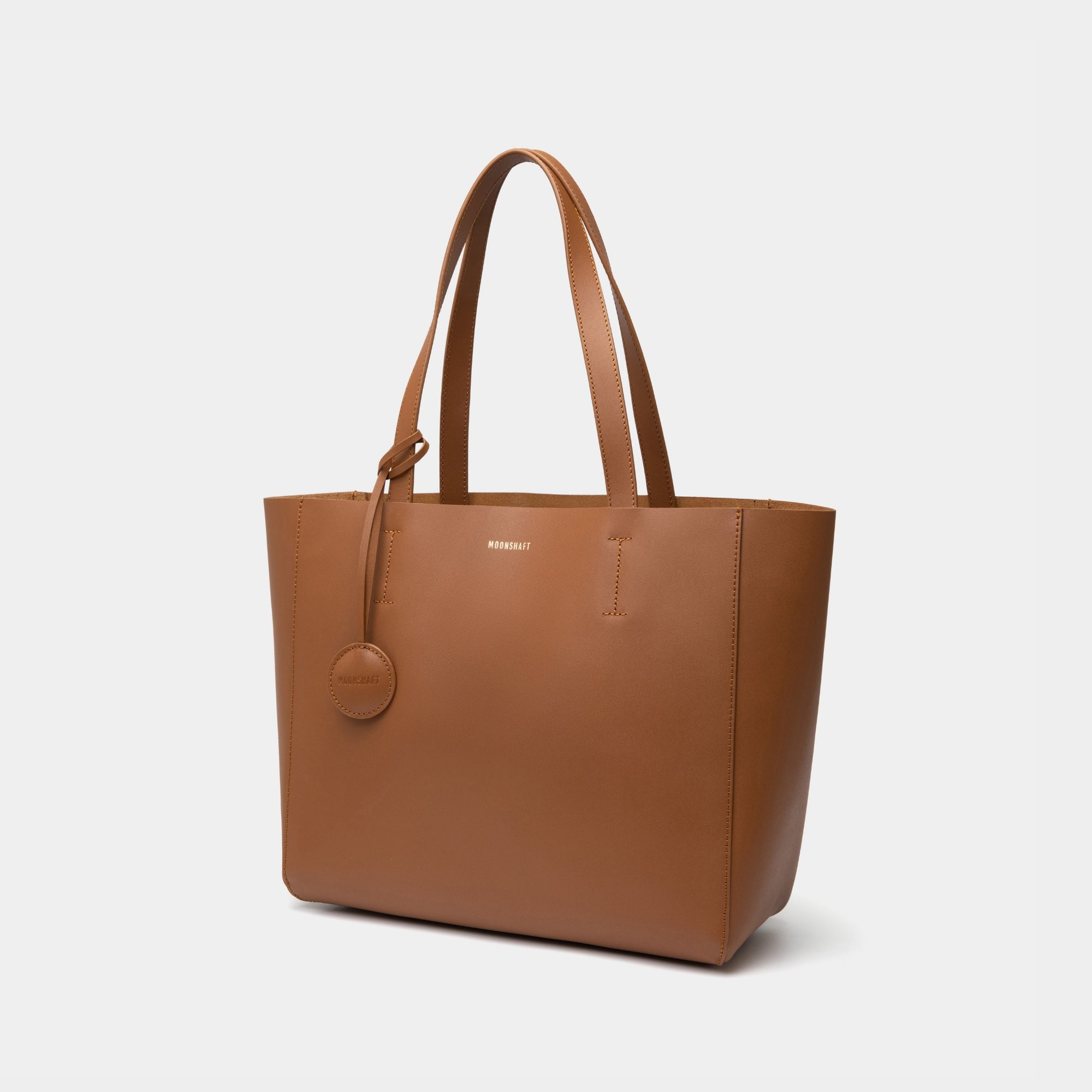 The diagonal view of the caramel MOONSHAFT tote bag displayed against a white background. The bag features a sleek, minimalist design with reinforced stitching along the handles, emphasizing its spacious and structured silhouette.