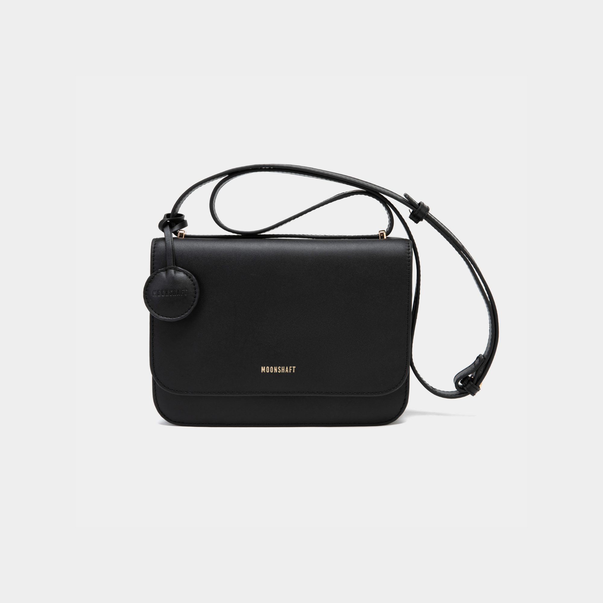 A stylish black-colored boxy leather handbag with a sleek, structured design. It features a circular leather tag attached near the top of the bag. The bag has clean lines and a minimalist aesthetic, with a long adjustable shoulder strap for versatile wear. The MOONSHAFT logo is elegantly embossed in gold at the front.
