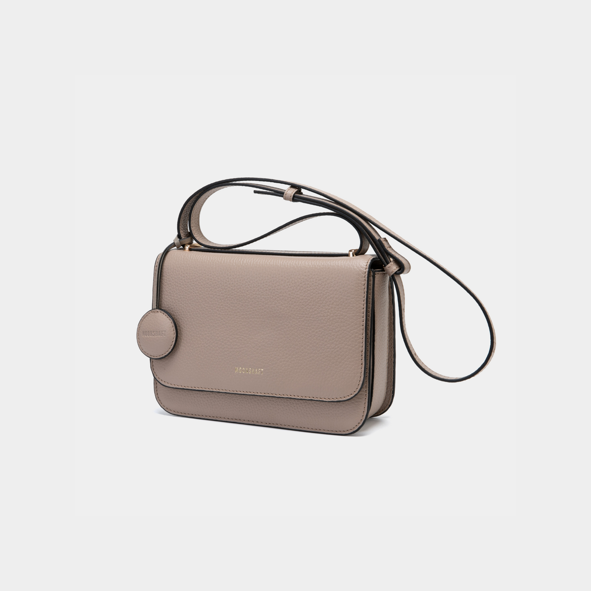 This image showcases a structured clay-colored boxy leather bag with a sleek, modern design. The bag features clean lines, a fold-over flap secured with a hidden closure, and a detachable circular leather tag on the side. The shoulder strap is adjustable, making it versatile for both crossbody and shoulder wear. 