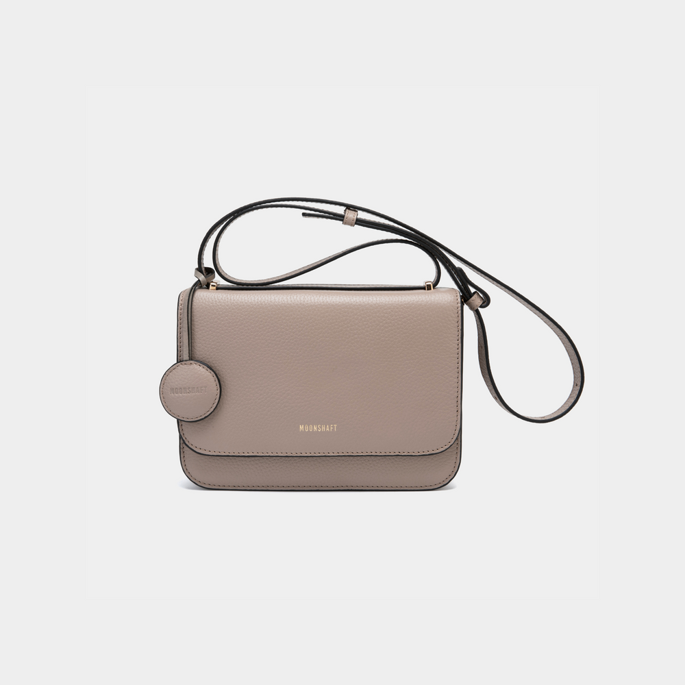 A stylish clay-colored boxy leather handbag with a sleek, structured design. It features a circular leather tag attached near the top of the bag. The bag has clean lines and a minimalist aesthetic, with a long adjustable shoulder strap for versatile wear. The MOONSHAFT logo is elegantly embossed in gold at the front.