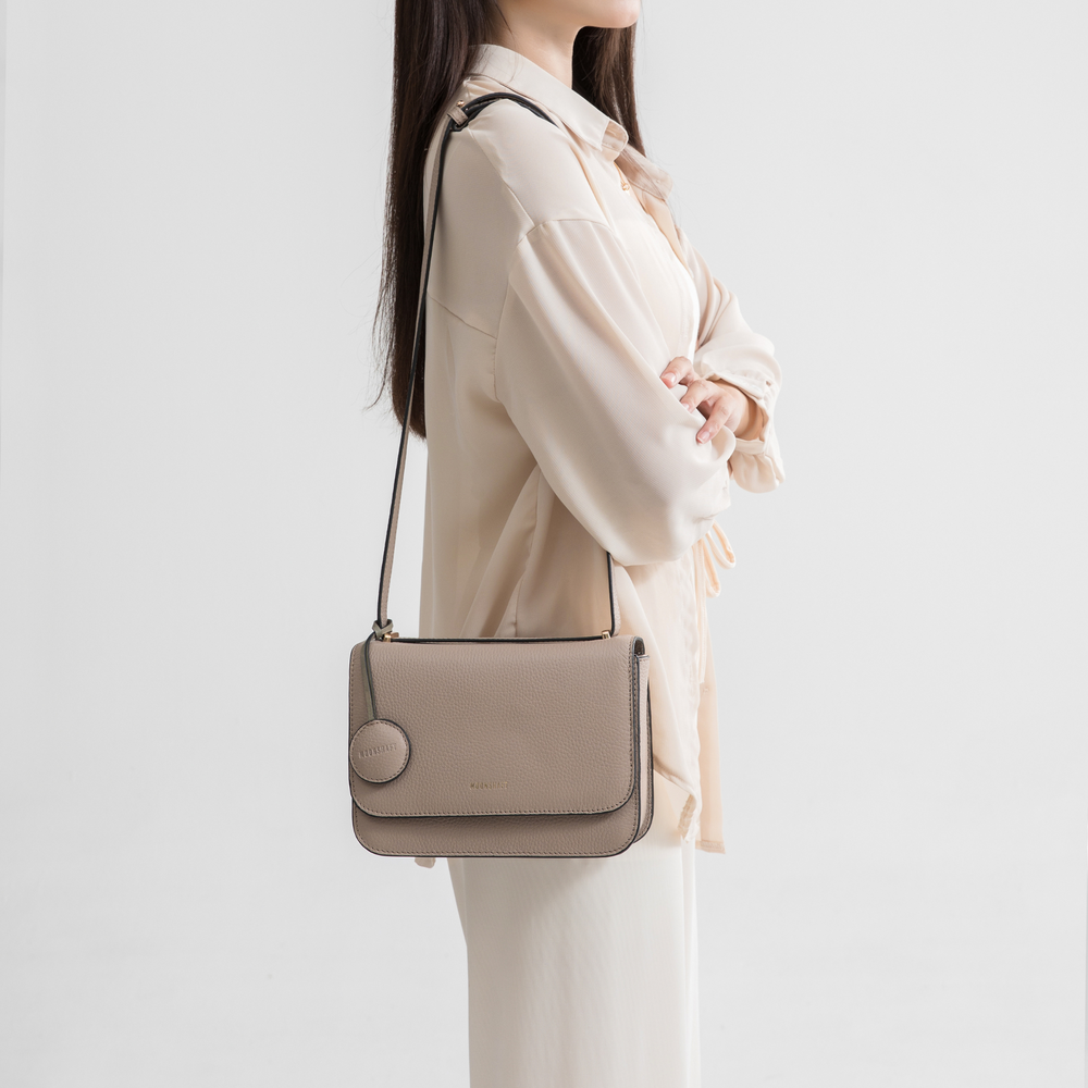 A woman in a light, neutral-colored outfit carries a clay-colored boxy leather bag on her shoulder. The bag has a structured, rectangular shape with an adjustable shoulder strap, making it easy to wear crossbody or over the shoulder. A circular leather tag is attached near the top, and the MOONSHAFT logo is embossed in gold at the front. The bag's minimalist design pairs well with her chic and elegant outfit, creating a sophisticated look.