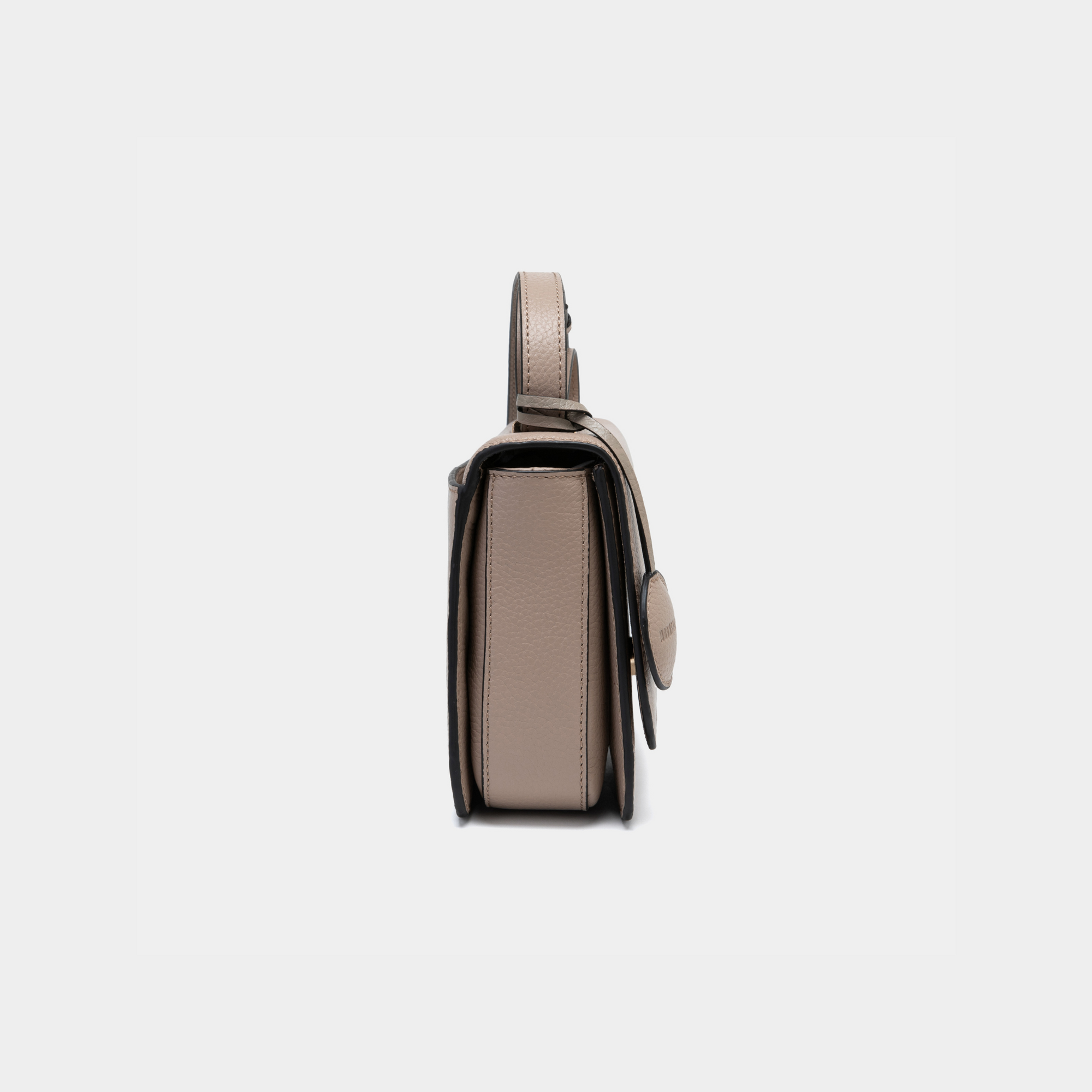 This side view of the boxy bag in clay color highlights its structured and rectangular shape. The smooth pebbled leather and minimal seams enhance the sleek silhouette, and the slim profile makes it appear compact. A circular leather tag is attached to the side, adding a subtle design element without compromising the bag's clean lines. The edges are neatly stitched, showcasing the craftsmanship of the bag. The strap attachment at the top is visible, blending seamlessly into the overall design.