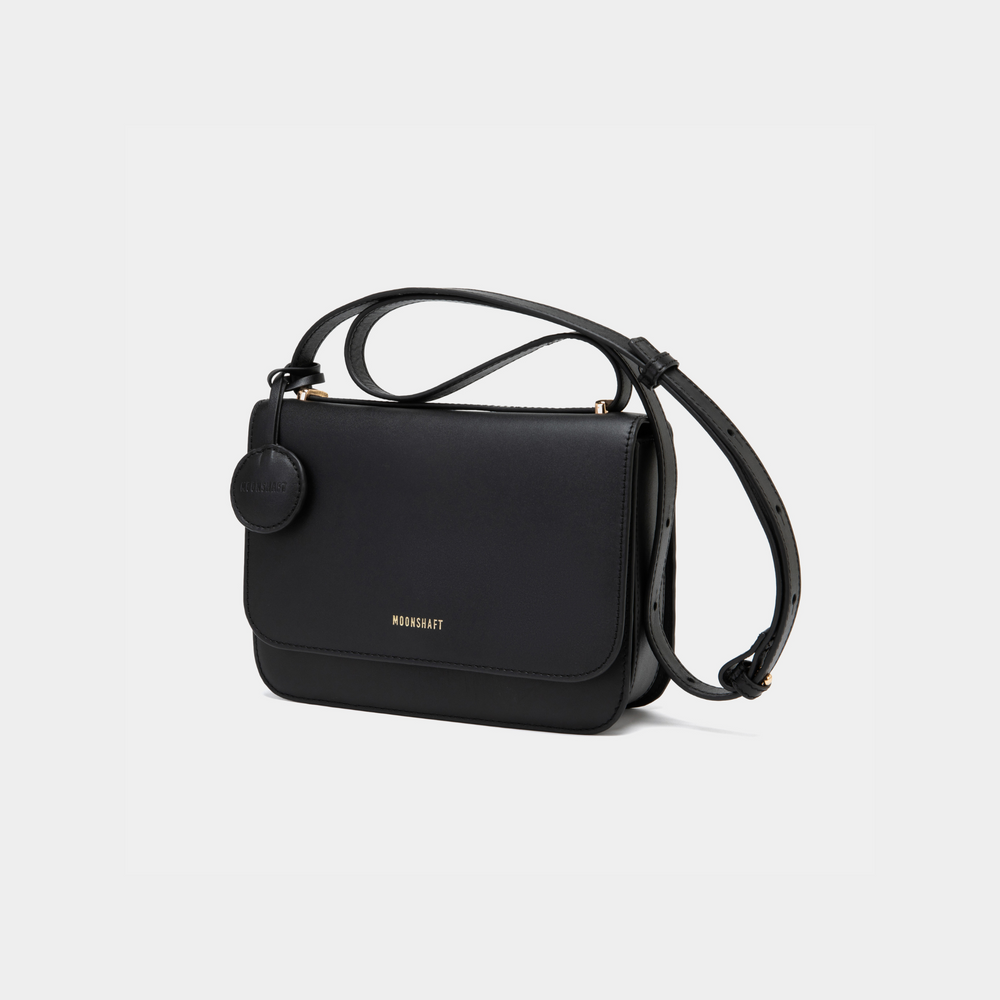  This image showcases a structured black-colored boxy leather bag with a sleek, modern design. The bag features clean lines, a fold-over flap secured with a hidden closure, and a detachable circular leather tag on the side. The shoulder strap is adjustable, making it versatile for both crossbody and shoulder wear. 