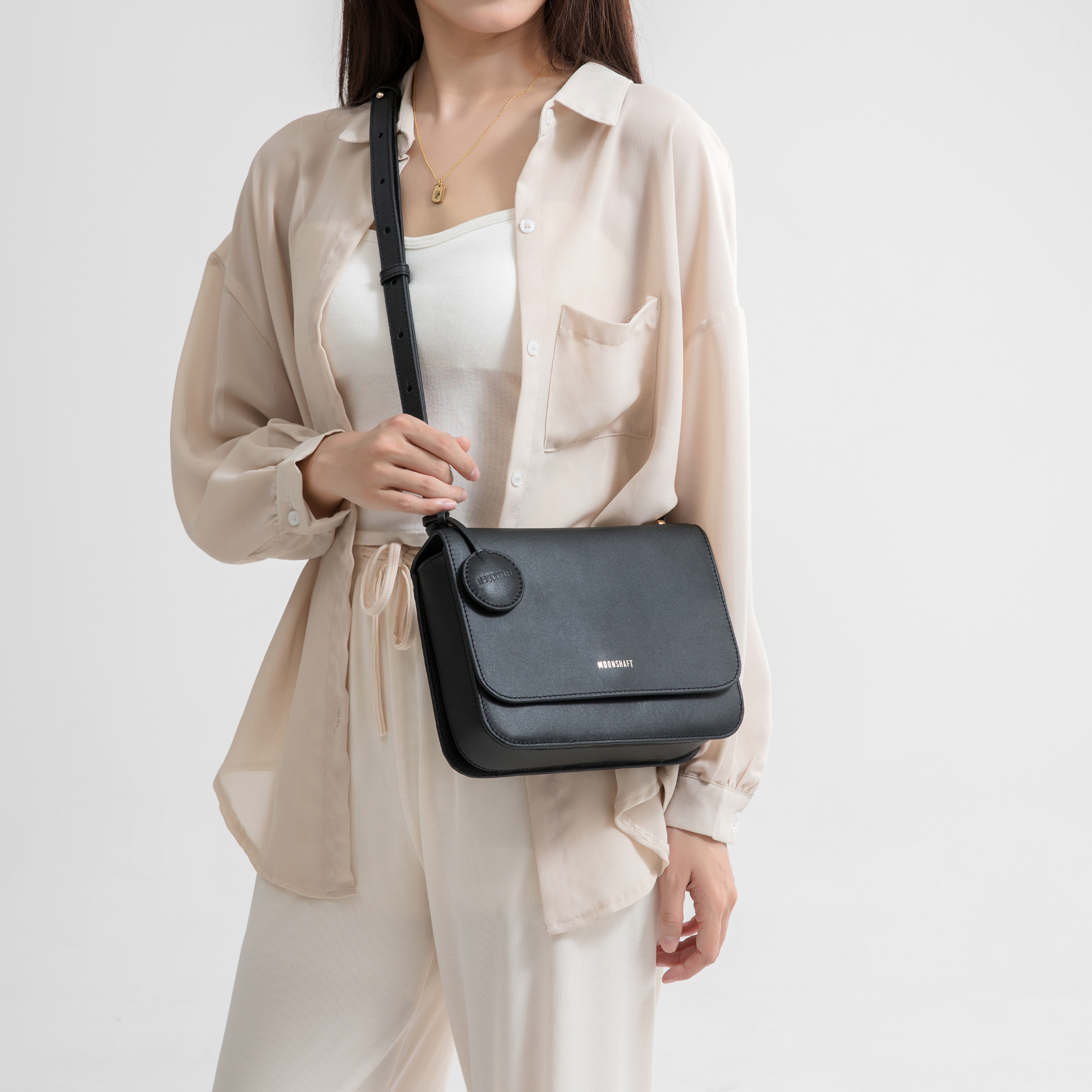 The model is wearing a black boxy bag with a smooth, structured shape. The bag features a slim, adjustable strap worn crossbody, making it versatile for everyday use. A circular leather tag is attached to the side, adding a simple yet elegant accent. The clean and minimalist design is enhanced by the subtle MOONSHAFT logo embossed on the front. The outfit, consisting of a light, flowing shirt and relaxed trousers, contrasts with the bag’s sleek black leather, creating a sophisticated and effortless look.