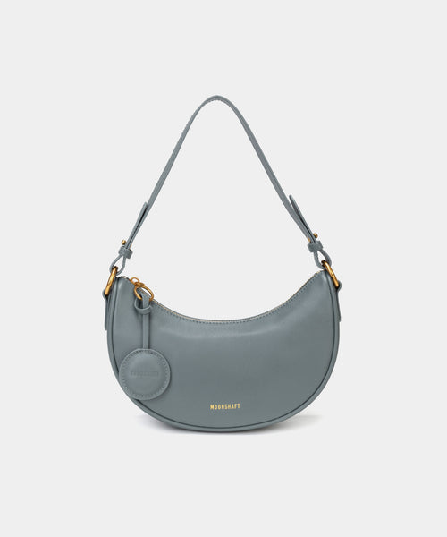 Front view of the Crescent Moon bag in ash blue, showcasing its smooth leather finish, curved silhouette, and gold hardware. The top handle is visible, along with an attached round coin purse accessory on the side. The minimal and elegant design highlights the bag's clean lines and structured shape.