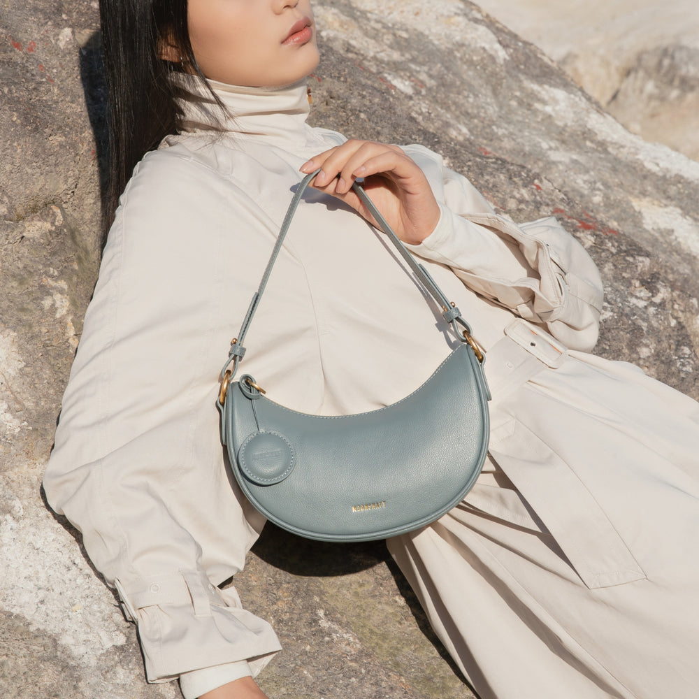 A model dressed in a light beige outfit lounges against a stone backdrop, holding the Crescent Moon bag in ash blue. The smooth leather bag, with its gold-tone hardware and attached coin pouch, rests elegantly on her lap. The bag's soft, crescent shape complements the minimalist and relaxed styling, while the MOONSHAFT logo in gold adds a touch of luxury. The natural outdoor setting, combined with the muted tones of the model's attire and the bag, creates a serene and sophisticated look.