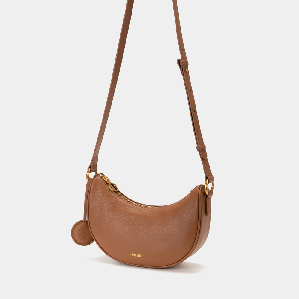 Front view of the Crescent Moon bag in caramel color, featuring smooth leather, gold-tone zipper closure, and a detachable adjustable strap. The bag's sleek, curved silhouette showcases its minimalist yet functional design, suitable for both casual and formal outfits.