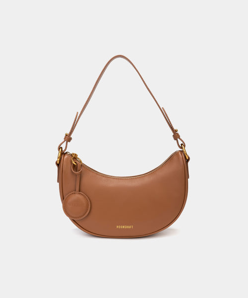 Front view of the Crescent Moon bag in caramel, showcasing its smooth leather finish, curved silhouette, and gold hardware. The top handle is visible, along with an attached round coin purse accessory on the side. The minimal and elegant design highlights the bag's clean lines and structured shape.