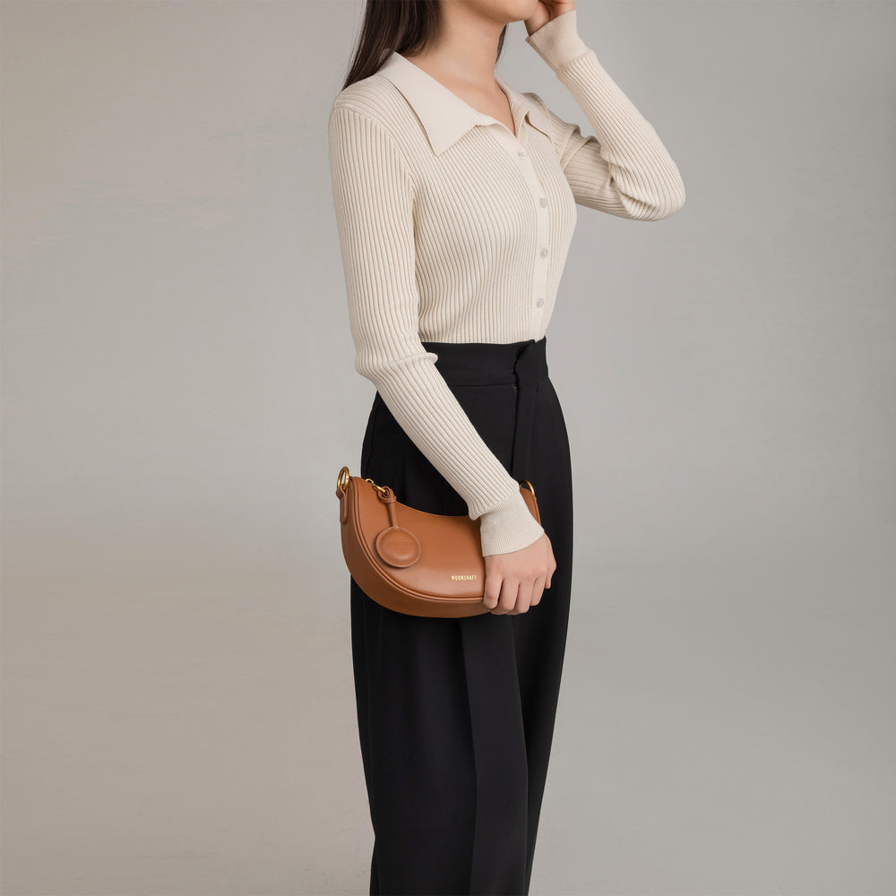 Model holding the Crescent Moon bag in caramel smooth leather at her side, wearing a beige ribbed button-up top paired with black pleated trousers. The sleek, minimalist design of the bag is highlighted by its compact size, curved shape, and gold hardware, making it a versatile accessory for both casual and formal outfits.