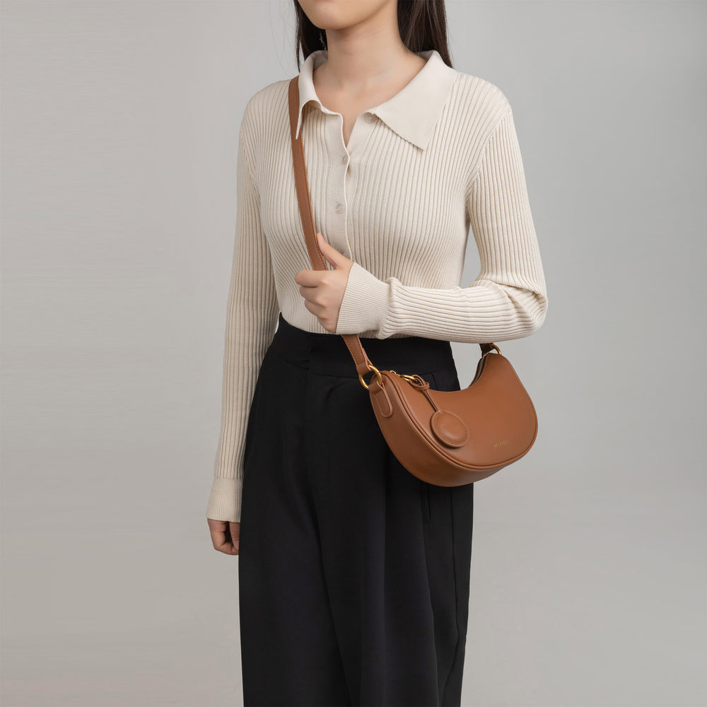 Model wearing the Crescent Moon bag in caramel smooth leather across her shoulder. The bag is worn with a longer strap, draped casually at the hip. The model wears a beige ribbed long-sleeve top with a collar, paired with black high-waisted pleated trousers, creating a chic and elegant look.