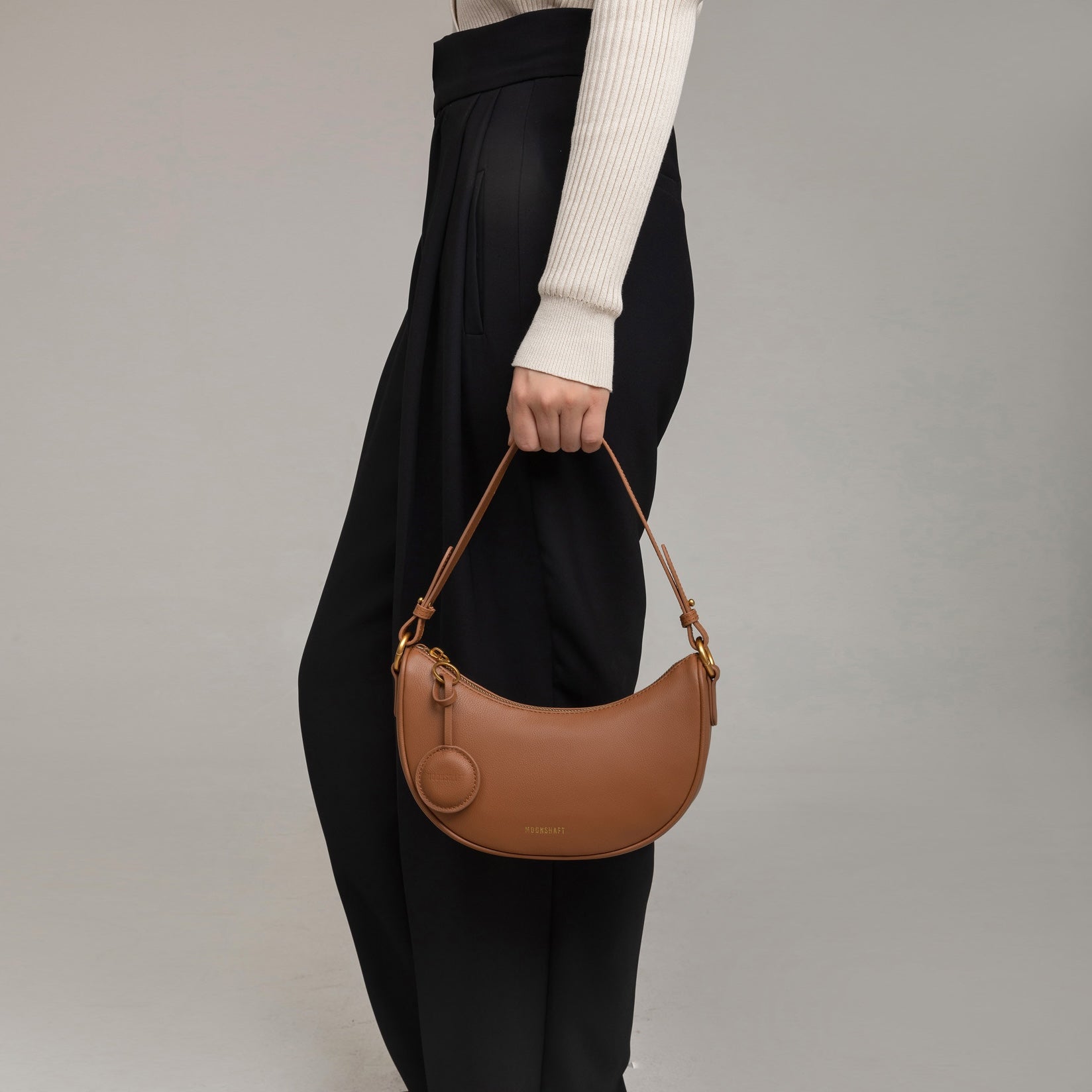 Close-up of a model holding the Crescent Moon bag in caramel smooth leather by her side. The bag is carried by its short handle, showcasing its compact and curved shape. The model wears black high-waisted trousers with pleats, paired with a beige ribbed long-sleeve top, complementing the bag's sophisticated and minimalist design.