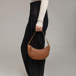 Close-up of a model holding the Crescent Moon bag in caramel smooth leather by her side. The bag is carried by its short handle, showcasing its compact and curved shape. The model wears black high-waisted trousers with pleats, paired with a beige ribbed long-sleeve top, complementing the bag's sophisticated and minimalist design.