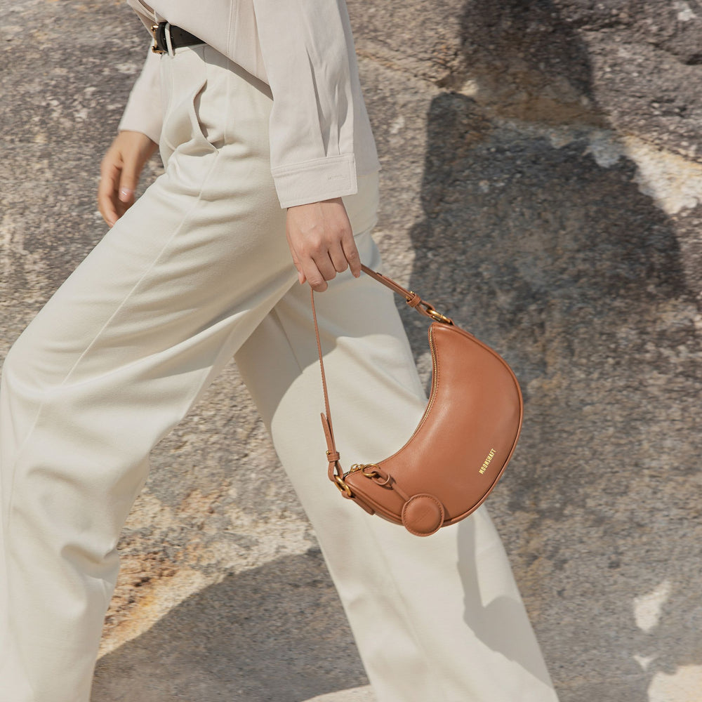 Close-up of a model walking while holding the Crescent Moon bag in caramel by the handle, paired with light beige pants. The smooth leather finish of the bag complements the neutral tones, adding a warm contrast to the outfit.