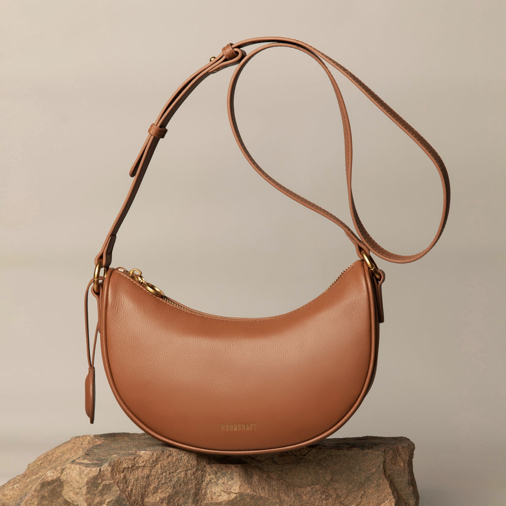Caramel smooth leather Crescent Moon bag displayed on a textured rock surface. The bag features an adjustable long strap with gold-tone hardware and a minimalist design. Its clean, curved silhouette is accentuated by the natural background, creating a refined, elegant aesthetic.